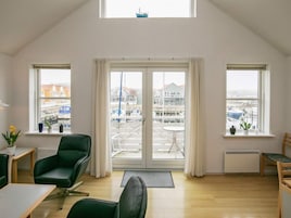 Una habitación con techos altos, dos ventanas con vistas a un puerto deportivo, una puerta corrediza de vidrio, una silla verde, una mesita pequeña y una planta en maceta.