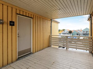 A wooden balcony with a white railing, a door with a number sign, and a view of buildings in the background.