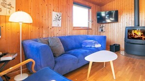 A cozy cabin interior with a blue sofa, a white table, a TV, and a wood-burning stove. - 6 Person Holiday Home in Humble (Sydlangeland)