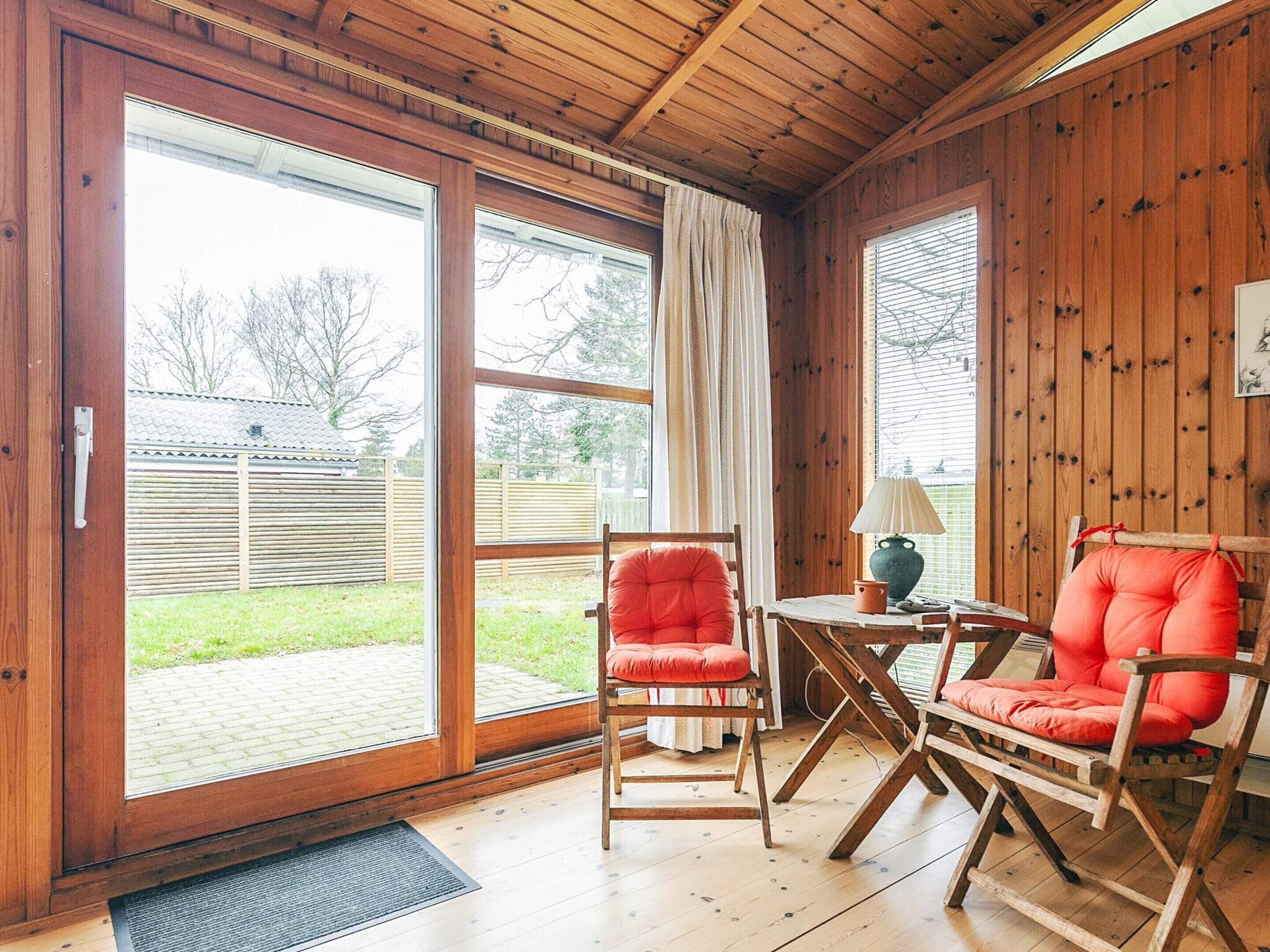 A wooden interior with a sliding glass door leading to a backyard, a red cushioned chair, and a small table with a lamp.