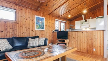 A cozy cabin interior with a wooden ceiling, a black leather sofa, a dining table with a unique design, a television, and a kitchenette.
