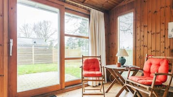 A wooden interior with a sliding glass door leading to a backyard, a red cushioned chair, and a small table with a lamp.