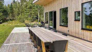 A modern wooden deck with a long table and chairs, surrounded by a forest.