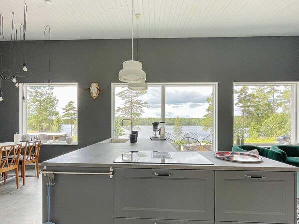 A modern kitchen with a large island, two windows with a lake view, and a ceiling with exposed beams. - 5 Star Holiday Home in Hacksvik (Håcksvik)