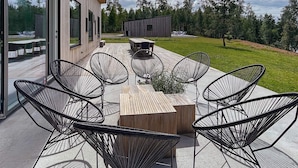 Ein Außenbereich mit modernen Stühlen und einem Holztisch, umgeben von Glaswänden und Blick auf eine bewaldete Landschaft.