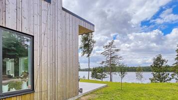 A modern wooden building with a large window, a concrete deck, and a view of a lake with trees.