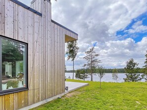 A modern wooden building with a large window, a concrete deck, and a view of a lake with trees. - 5 Star Holiday Home in Hacksvik (Håcksvik)