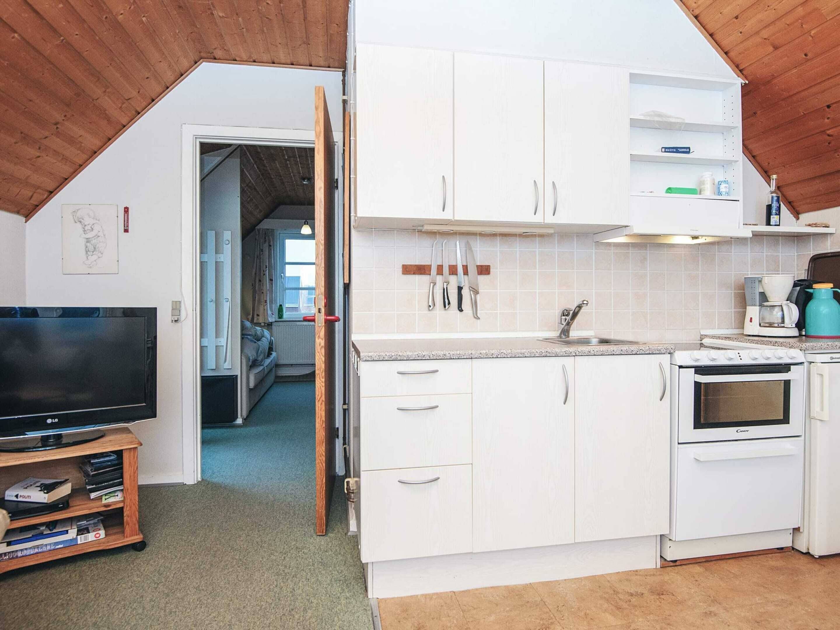 Una cocina compacta con gabinetes blancos, horno integrado y lavamanos. Hay un televisor sobre un mueble bajo y una estantería con libros.