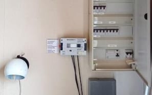 Un tableau électrique fixé au mur, doté d’un panneau de commande et d’un appareil blanc, possiblement un thermostat, le tout relié par des câbles.