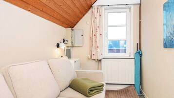 A small, cozy room with a wooden ceiling, a white radiator, a window with floral curtains, a white sofa with a green throw, and a blue towel on the floor.