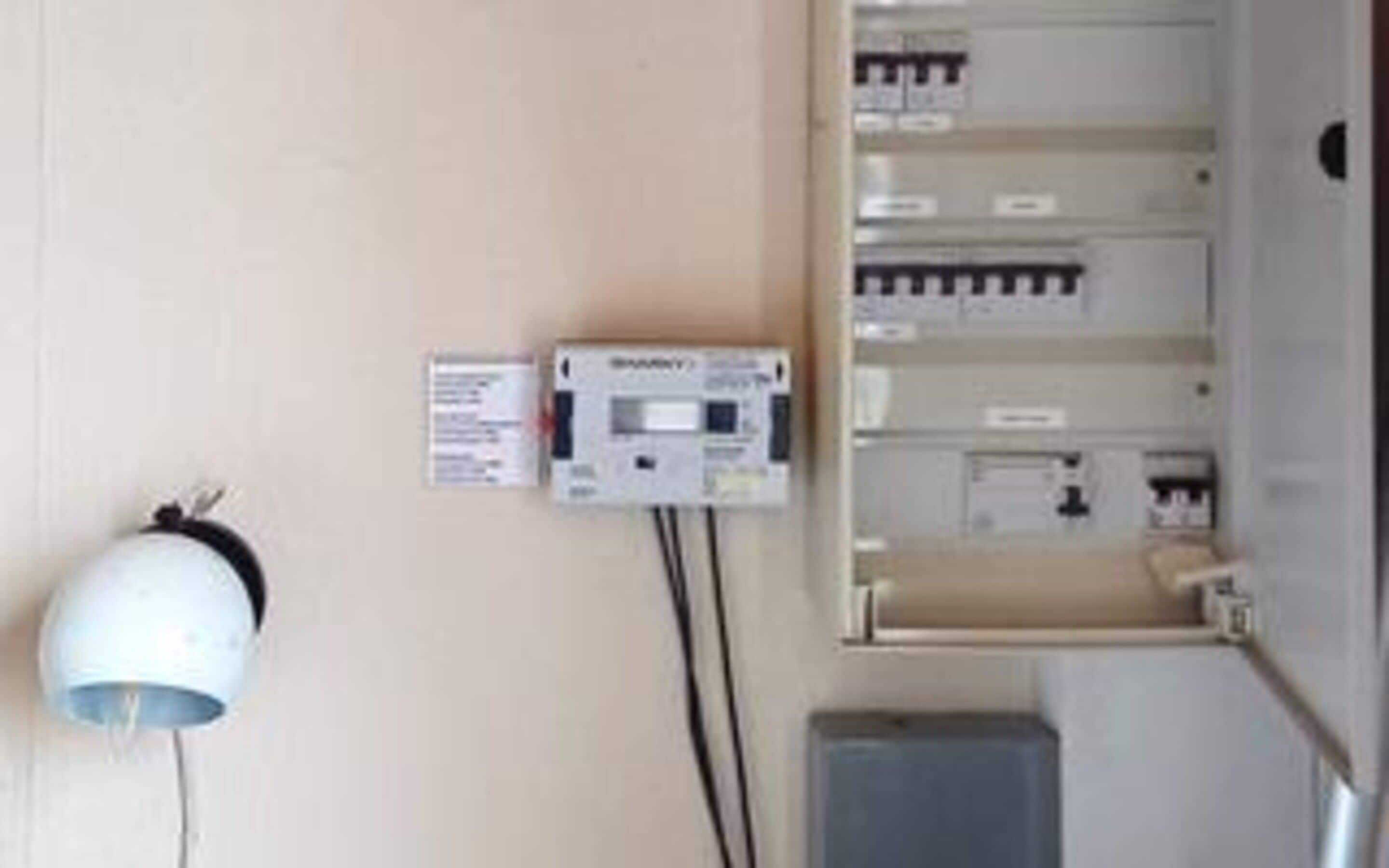 Een wandpaneel met elektrische installatie, een bedieningspaneel en een wit apparaat, mogelijk een thermostaat, verbonden door bedrading.