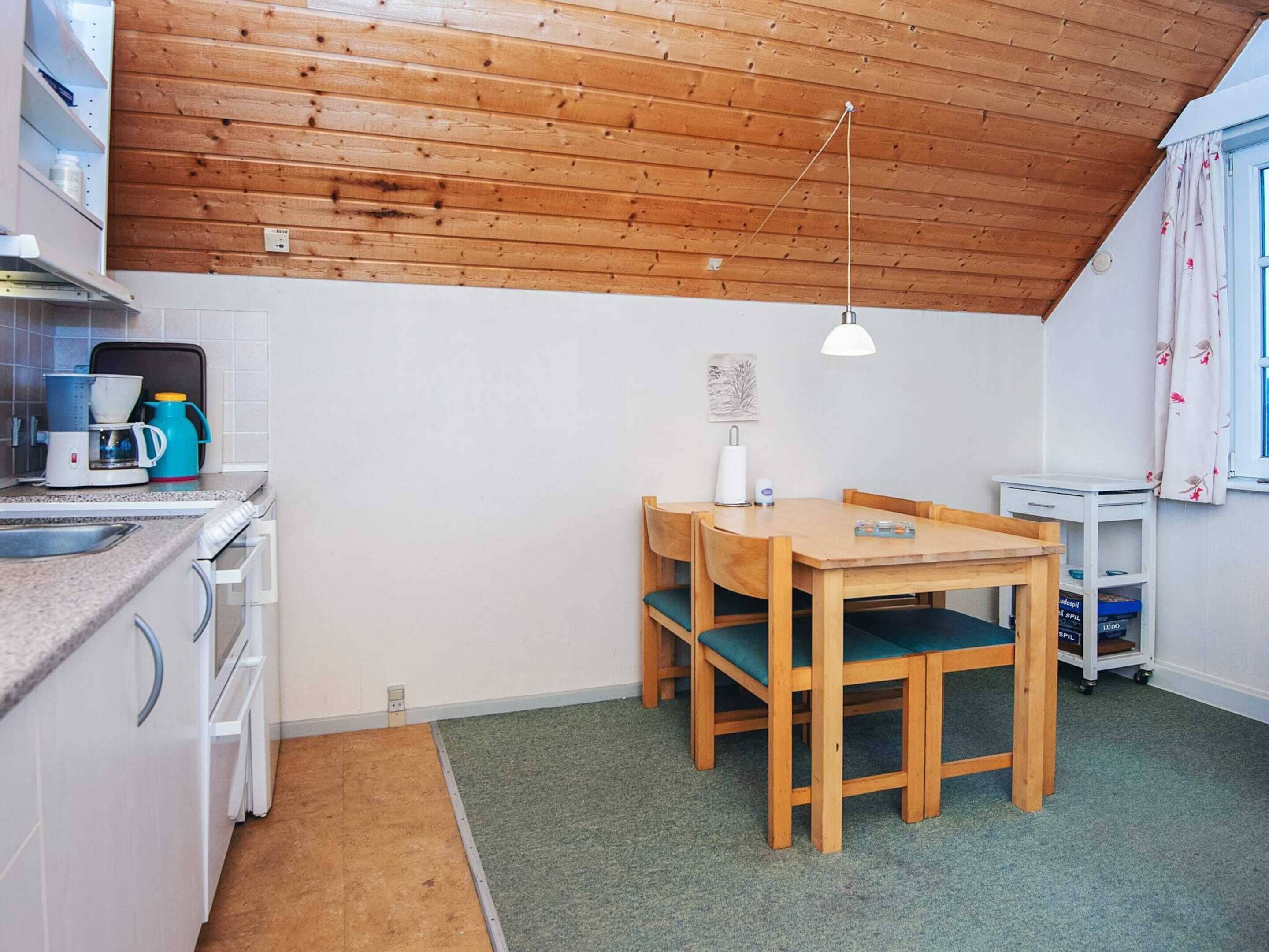 A compact kitchen with wooden ceiling, a dining table with chairs, a coffee maker, and a small shelf with books.