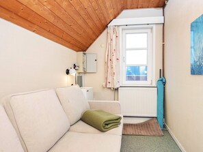 A small, cozy room with a wooden ceiling, a white radiator, a window with floral curtains, a white sofa with a green throw, and a blue towel on the floor. - 4 Person Holiday Home in Romo (Rømø)