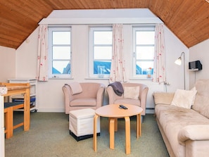 A cozy living room with a wooden ceiling, two windows with floral curtains, a beige sofa, a small round table with a remote control, and a wooden side table with books. - 4 Person Holiday Home in Romo (Rømø)