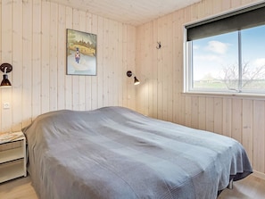 Quarto com cama, mesa de cabeceira, luminária na parede, quadro e janela.