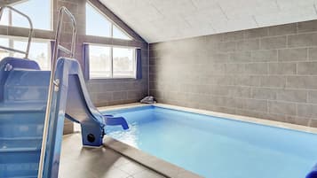 An indoor swimming pool with a slide, a ladder, and a view of the outside through a window.