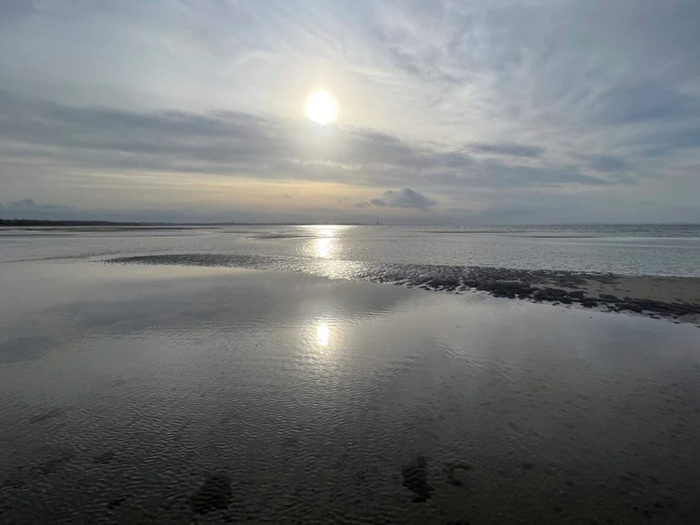 Une plage aux eaux calmes où se reflète le soleil, un ciel dégagé et une terre lointaine à l’horizon.