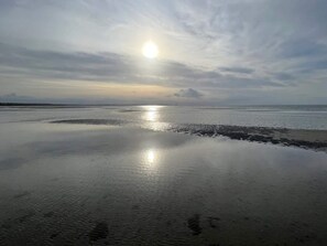 Una playa con aguas tranquilas que reflejan el sol, un cielo despejado y tierra a lo lejos.