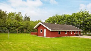 Un bâtiment rouge avec un toit en pente, une porte et des fenêtres, situé dans une zone herbeuse avec des arbres en arrière-plan.