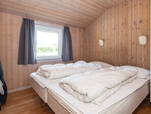 A bedroom with a wooden ceiling, a double bed with white bedding, a window with curtains, and a wall-mounted light fixture.