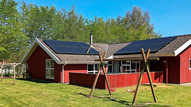 Un bâtiment rouge doté de panneaux solaires, une aire de jeux avec des balançoires et une cour couverte d’herbe.