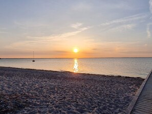 A beach with a wooden boardwalk leading to the water, a sailboat in the distance, and the sun setting over the horizon.