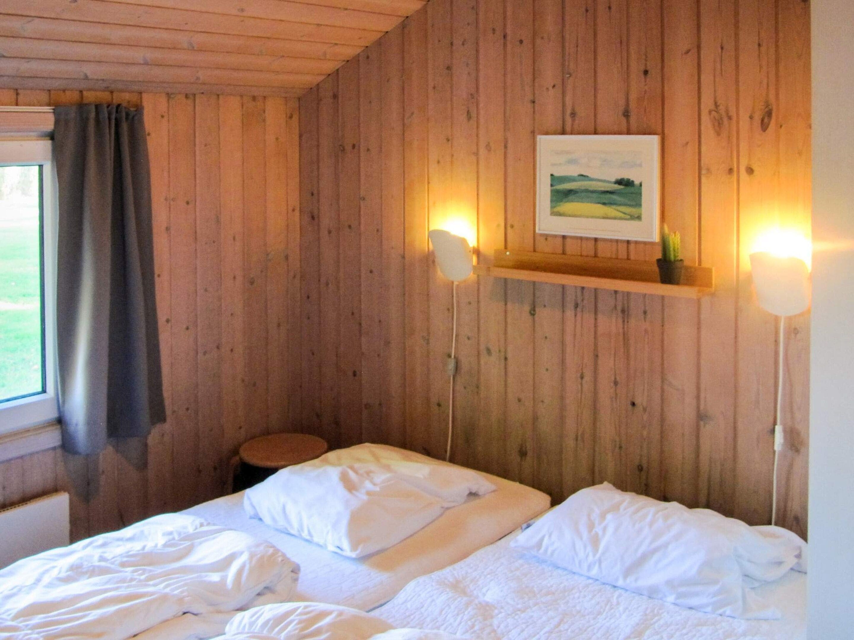 Una stanza dalle pareti in legno, con due letti con biancheria bianca, uno sgabello, una finestra con tenda e uno scaffale con una cornice e una pianta.