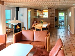 A cozy living room with a fireplace, a dining area with a wooden table and chairs, and a kitchen area with a stove and hanging lights. - 5 Star Holiday Home in Nordborg (Nordborg)