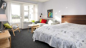 Ett sovrum med en stor säng, ett skrivbord, en stol, ett litet bord med en växt och ett fönster med gardiner.