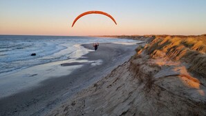 Une plage où l’on voit une personne faire du parapente, des dunes de sable et un ciel dégagé.