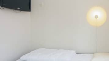 Una cama individual con ropa de cama blanca, un televisor montado en la pared y una lámpara fijada a la pared.