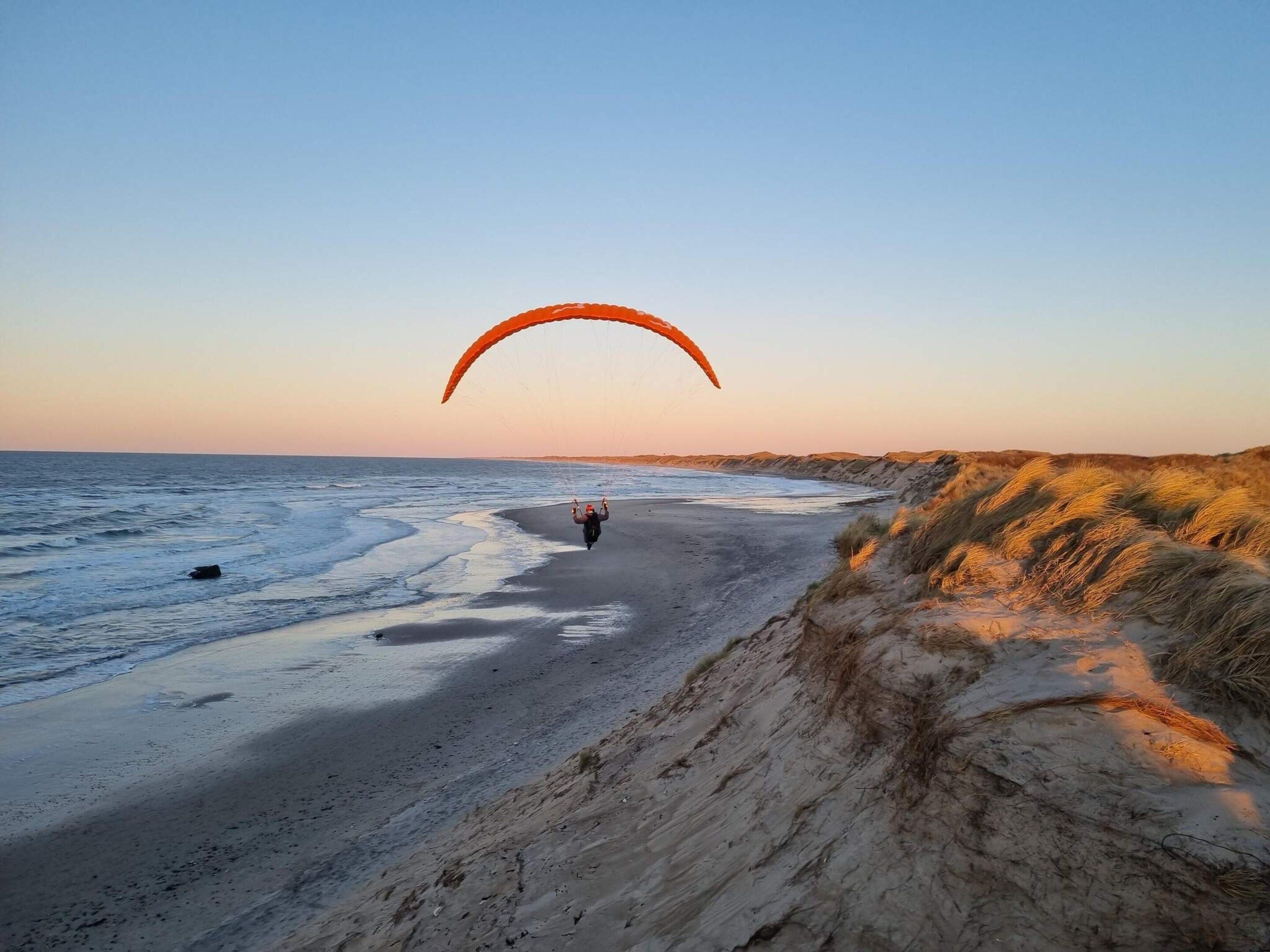 A beach with a person paragliding, sandy dunes, and a clear sky.