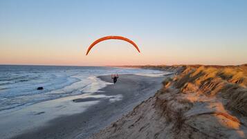 A beach with a person paragliding, sandy dunes, and a clear sky.