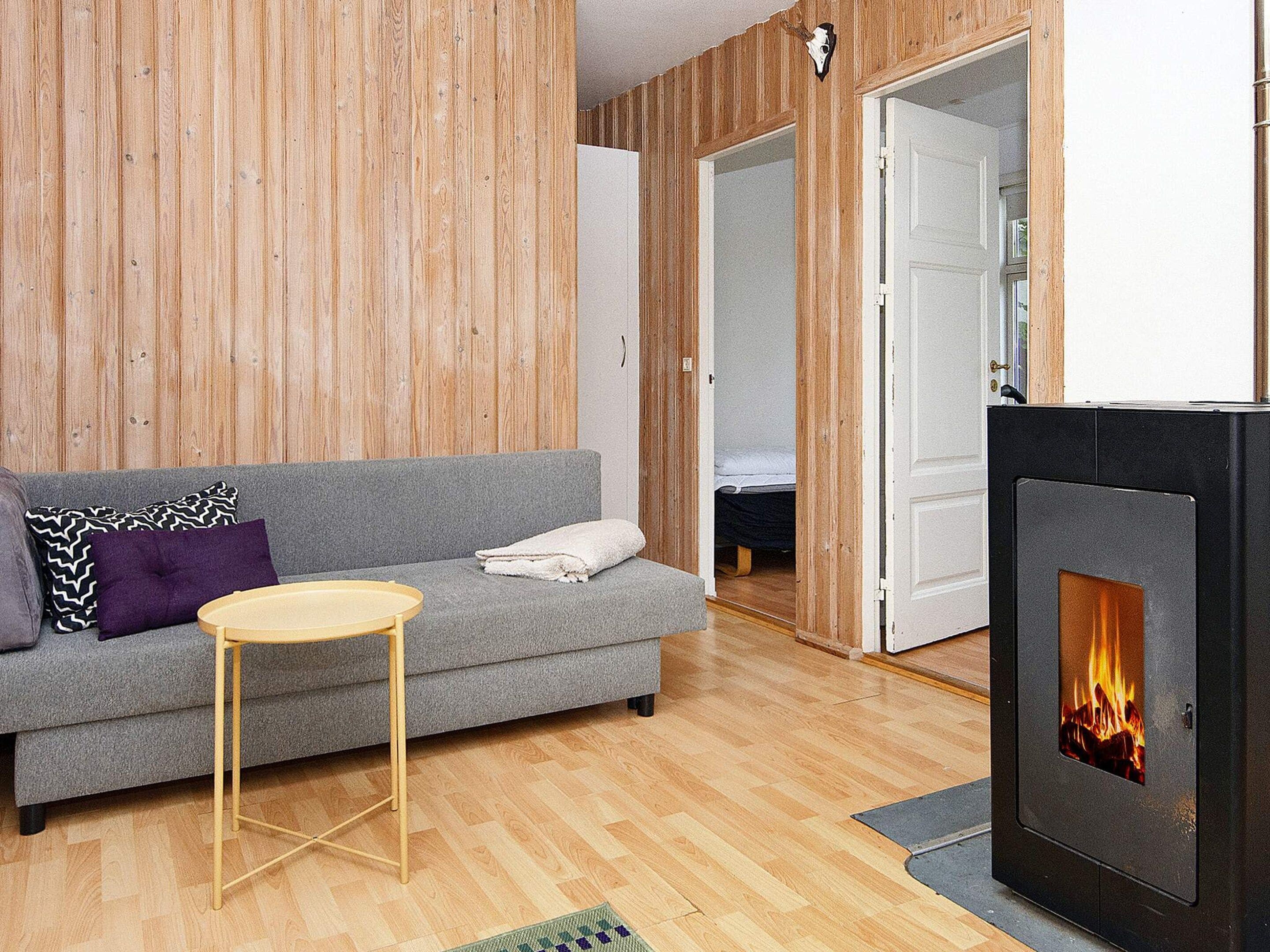 A modern living room with a grey sofa, a round wooden side table, a fireplace with visible flames, and wooden paneling on the walls.