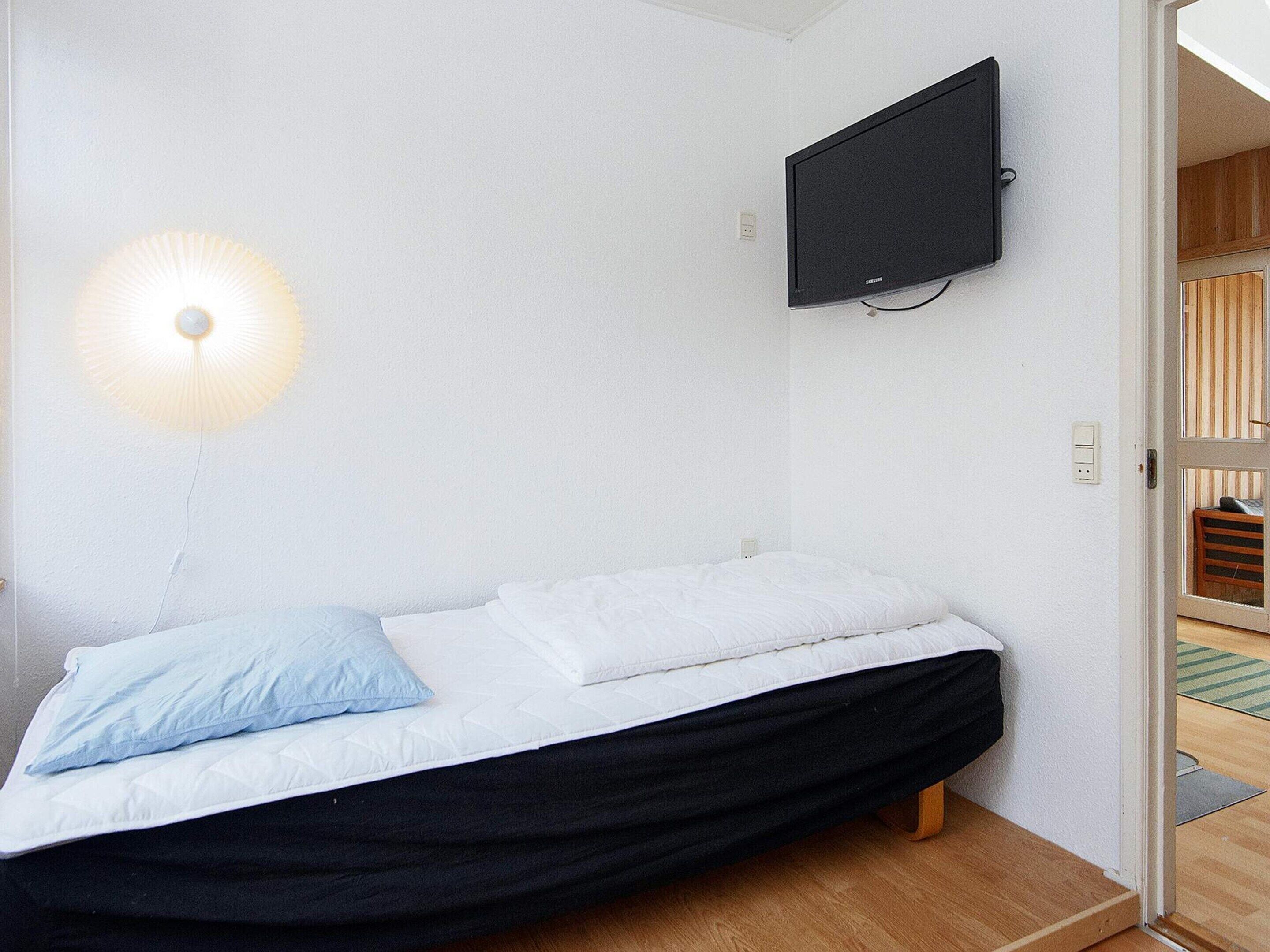 Una cama individual con estructura negra y ropa de cama blanca, un televisor montado en la pared y una lámpara a la izquierda.