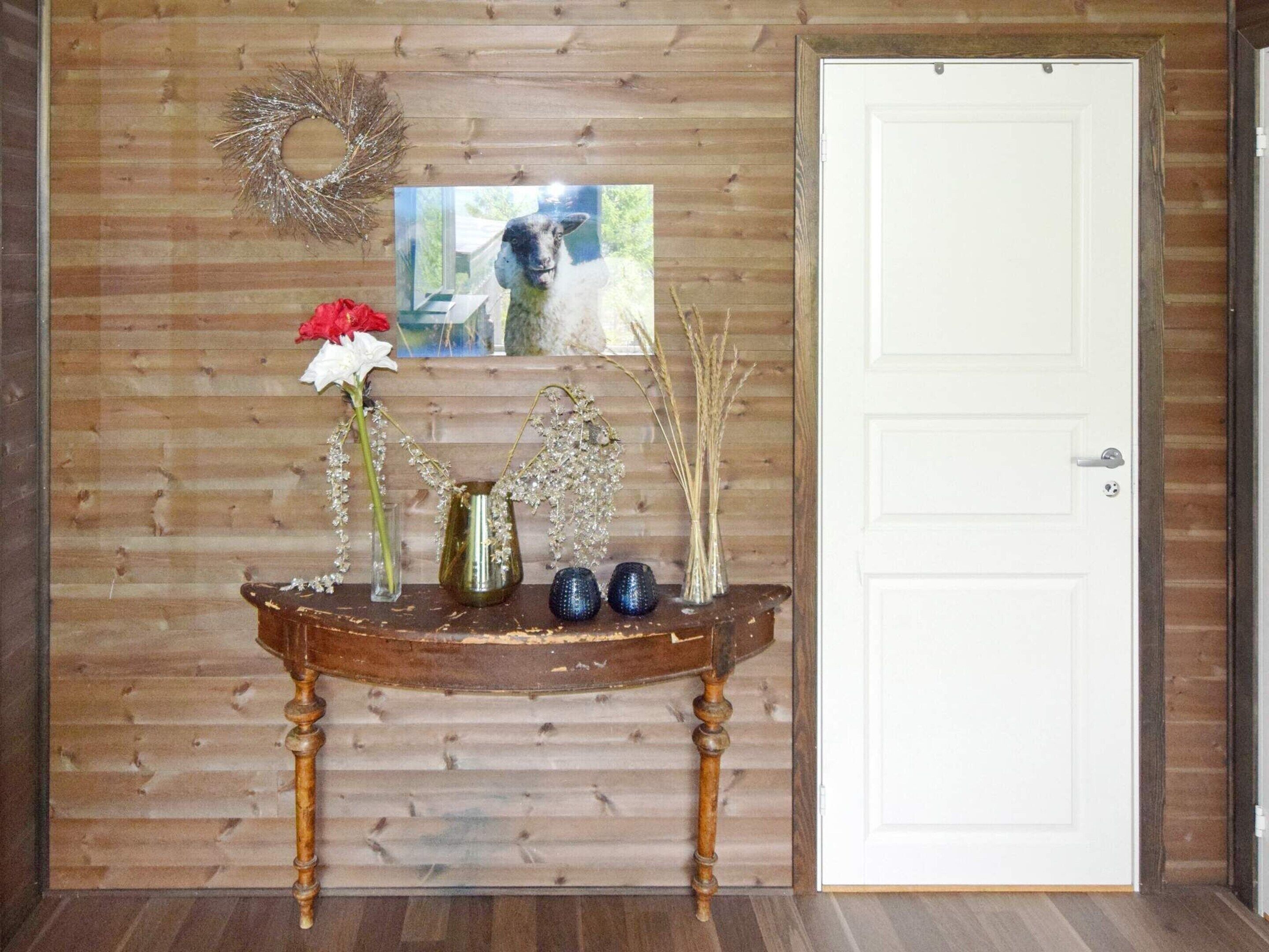 Una pared de madera con una puerta blanca, un arreglo decorativo, un cuadro enmarco y una pequeña mesa con jarrones y flores.
