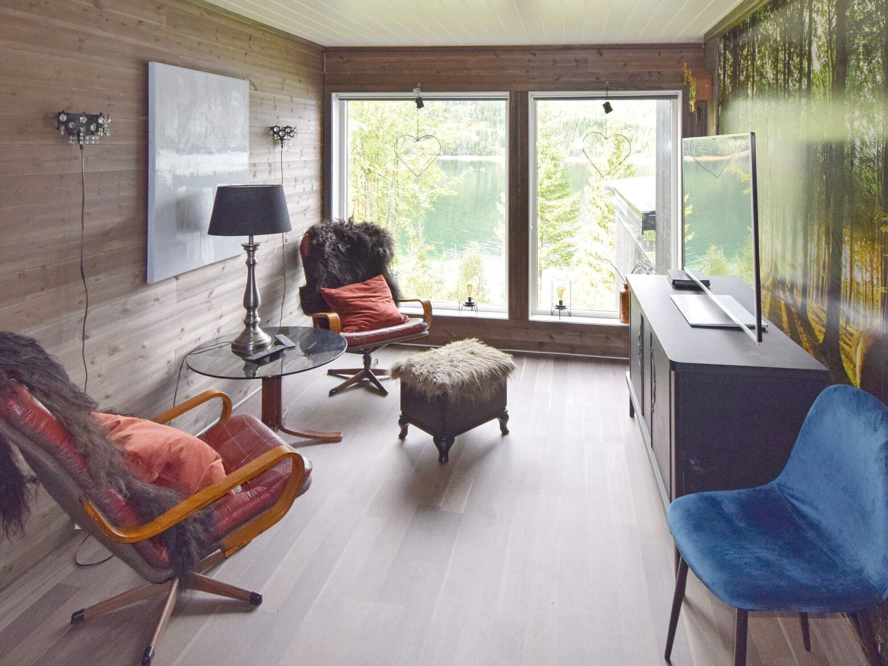 A cozy room with a wooden interior, a large window with a view of greenery, a desk with a computer, and a comfortable seating area with a chair and ottoman.
