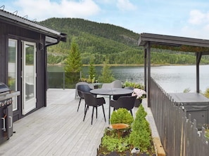 A deck with a grill, chairs, and a table overlooking a lake with a forested hill in the background. - 9 Person Holiday Home in Foldereid (Nærøysund)