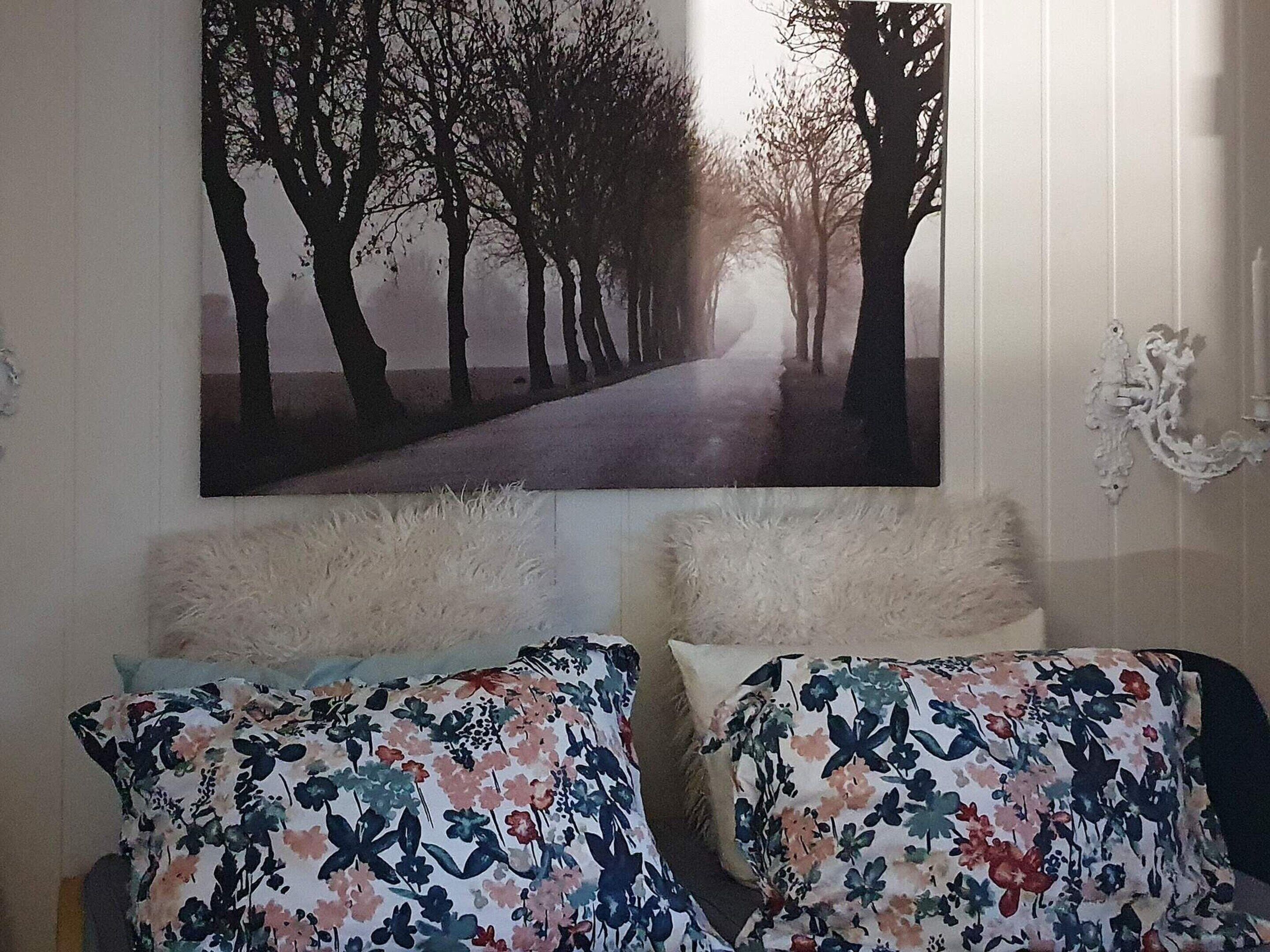 A bedroom with two beds, each with floral-patterned bedding, a large framed picture of a tree-lined path on the wall, and a decorative wall piece.