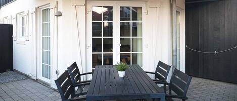 Un patio con una mesa, sillas, una planta en maceta y un ventanal corredera que da a un espacio interior.