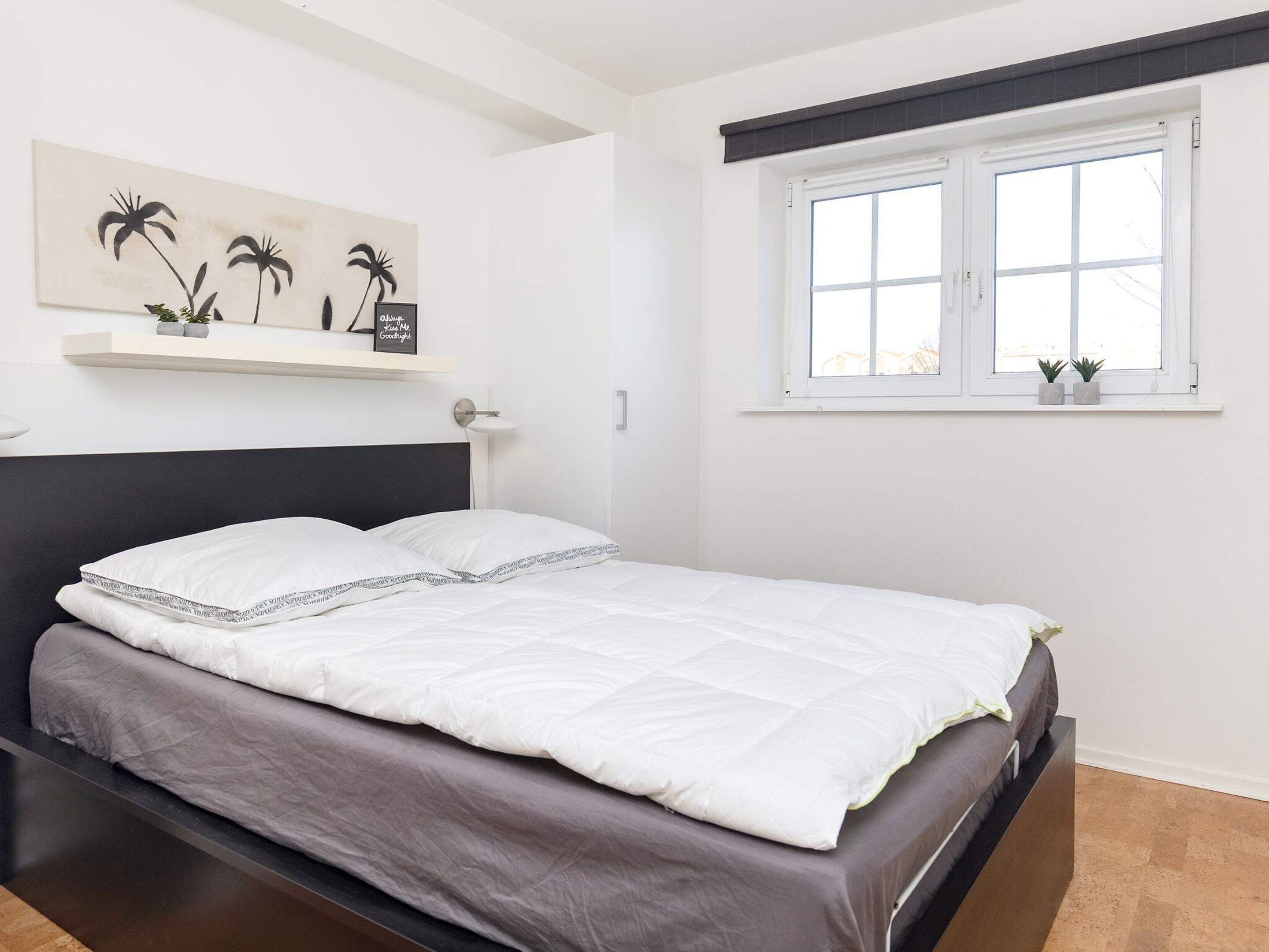 A bedroom with a bed, a window with blinds, a wall-mounted shelf with plants, and a painting.