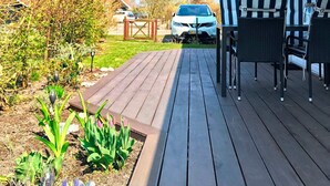 A wooden deck with outdoor seating, a garden with plants and flowers, and a parked car in the background. - 6 Person Holiday Home in Vaeggerlose (Vaeggerlose)
