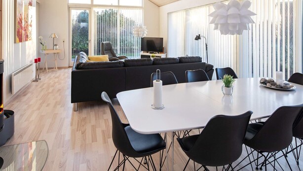 A modern living room with a black sofa, a dining table with chairs, and a large window with blinds. - 6 Person Holiday Home in Vaeggerlose (Vaeggerlose)