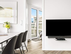 A modern living room with a flat-screen TV mounted on the wall, a dining area with a white table and black chairs, and a large window offering a view of the outside. - 5 Star Holiday Home in Wendtorf (Wendtorf)