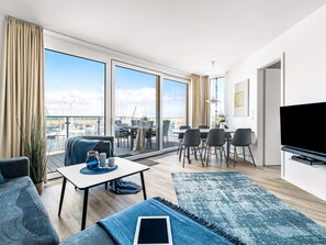 A modern living room with a large TV, a dining area, and a view of the marina. - 5 Star Holiday Home in Wendtorf (Wendtorf)