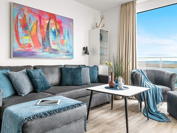 Un salon moderne avec un canapé gris, une table basse et une vue sur la mer grâce à de grandes fenêtres.