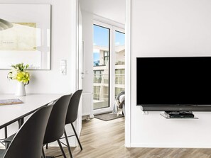 A modern living room with a flat-screen TV mounted on the wall, a dining area with a white table and black chairs, and a large window offering a view of the outside. - 5 Star Holiday Home in Wendtorf (Wendtorf)