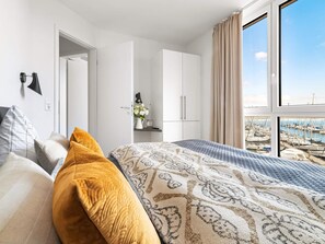 A hotel room with a bed, a desk, a chair, a lamp, and a view of a marina through large windows. - 5 Star Holiday Home in Wendtorf (Wendtorf)