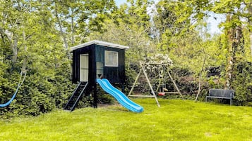 Casetta dei giochi con scivolo e altalena in un prato circondato da alberi.