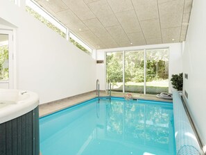An indoor swimming pool with a hot tub, large windows, and a view of greenery outside.